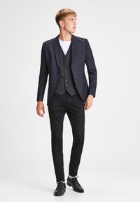 Traje de tres piezas negro con chaqueta ajustada, chaleco y pantalones entallados; combinado con una camiseta blanca y zapatos negros. Textura de tela suave.