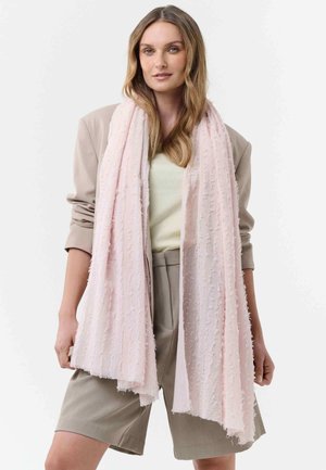 Woman with long wavy hair wears beige blazer and shorts, cream top, and light pink textured scarf draped around her neck.