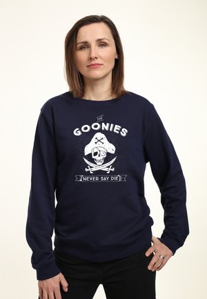 Donna con capelli castani fino alle spalle che indossa una felpa blu navy con una grafica di teschio e spade incrociate con la scritta "The Goonies Never Say Die".