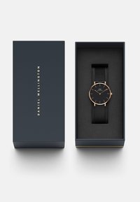 Black Daniel Wellington watch with a mesh strap, housed in a navy box. Features a rose gold case and minimalist black dial with thin markers.