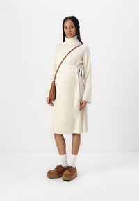 Rochie tricotată crem cu guler înalt, mâneci lungi, curea la talie și lungime până la jumătatea gambei. Asortată cu pantofi cu platformă maro și șosete albe.