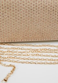 Chaîne en or disposée devant une pochette rose blush ornée de petits strass brillants.