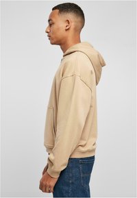Hoodie beige realizzato in misto cotone, con tasca frontale, vestibilità rilassata e cappuccio con coulisse. Indossato con jeans in denim blu.