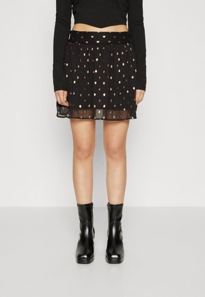 Frau trägt einen schwarzen Rock mit metallischen Punkten, ein schwarzes langärmliges Cropped-Top und schwarze Stiefeletten, steht vor einem schlichten Hintergrund.