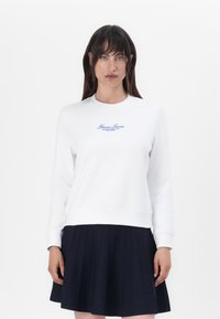 Felpa bianca in morbido tessuto, con logo ricamato blu sul petto, abbinata a una gonna a pieghe blu scuro.