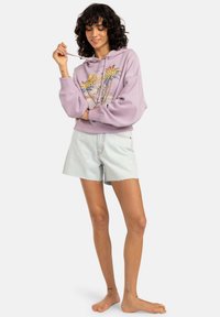Felpa lilla con design grafico e coulisse, abbinata a pantaloni shorts in denim chiaro con bordi sfilacciati. Modella in piedi a piedi nudi.