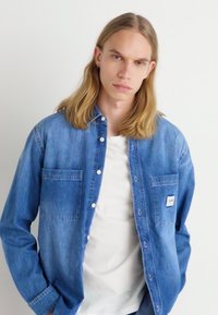 Jasnoniebieska koszula dżinsowa z zapięciem na guziki, dwiema kieszeniami na piersiach i białym przeszyciem. Nosi się ją na białej koszulce.