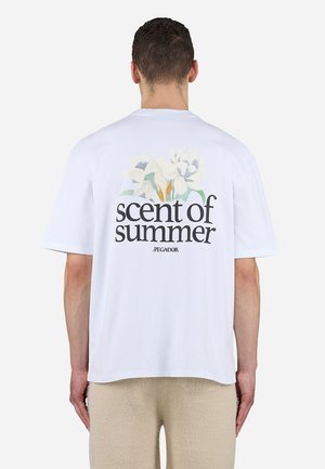Mann mit kurzen Haaren, der ein weißes T-Shirt mit dem Schriftzug „Duft des Sommers“ und floralem Design auf dem Rücken trägt, kombiniert mit beigen Hosen.