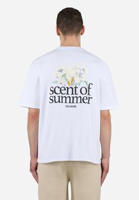 Uomo con capelli corti che indossa una maglietta bianca con la scritta "scent of summer" e un motivo floreale sulla schiena, abbinata a pantaloni beige.