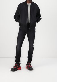 Chaqueta bomber negra con puños acanalados, suéter de punto gris, jeans oscuros y zapatillas negras con acentos rojos y suelas texturizadas.