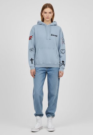 Haut à capuche bleu clair avec une poche avant, mettant en avant des motifs brodés, incluant des cœurs, une croix et du texte. Associé à un jean bleu et des baskets blanches.