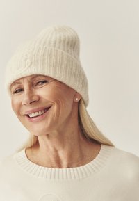 Caciula tricotată crem cu o textură cu nervuri, având un vârf lejer. Purtați cu un pulover ușor asortat, ambele în nuanțe moi și neutre.