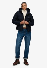 Sort pufferjakke med hætte, elastiske manchetter og et hvidt lynlogo. Sammen med lyseblå jeans og brune snørrebukser.