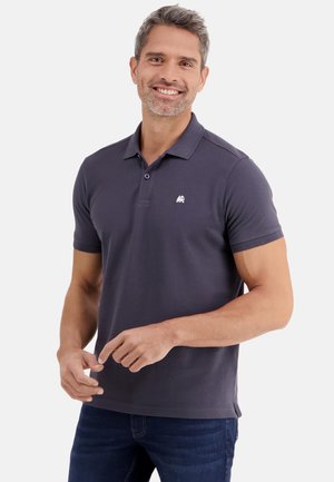 Lachende man met kort grijs haar, gekleed in een donkergrijs poloshirt en donkerblauwe spijkerbroek, staand tegen een effen witte achtergrond.