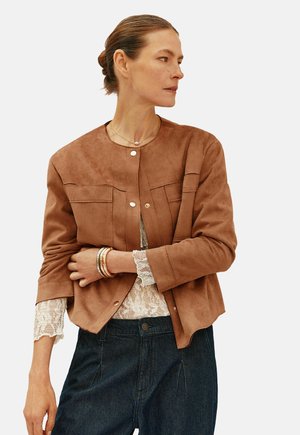 Giacca in suede marrone con scollo rotondo, chiusura frontale a bottoni e due tasche sul petto. Indossata sopra a un top di pizzo e abbinata a jeans scuri.