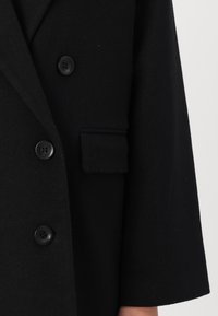 Svart ullfrakk med skreddersydd design, med store knapper, sidelomme og en teksturert overflate. Klassisk notch-lapel-detalj.