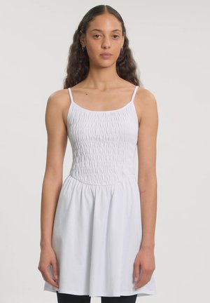 Junge Frau mit langen lockigen Haaren, die ein ärmelloses weißes Kleid mit einem enganliegenden, strukturierten Oberteil und einem ausgestellten Rock vor schlichtem Hintergrund trägt.
