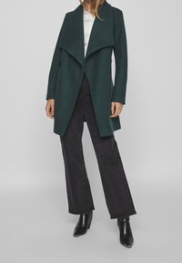 Tumma vihreä, avoin takki, jossa on roikkuva kaulus, valmistettu pehmeästä kankaasta. Yhdistettynä korkeavyötär öisiin mustiin leveälahkeisiin farkkuihin ja mustiin korkokenkiin.