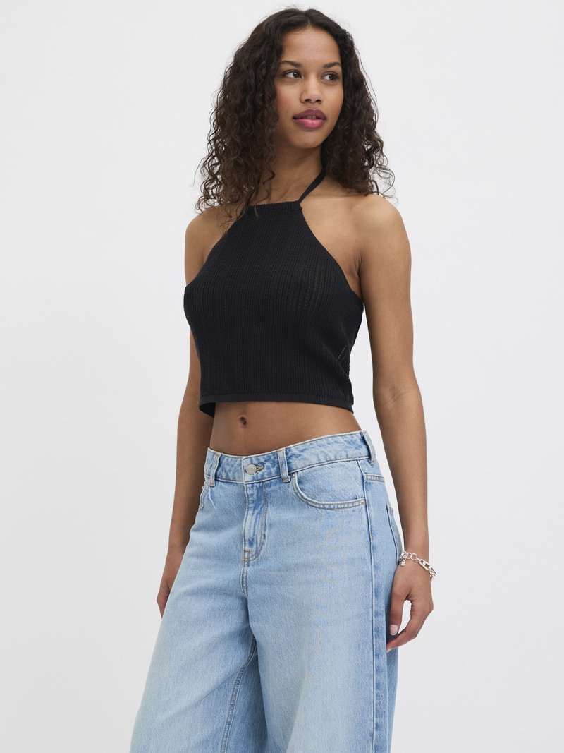 Ung kvinne med krøllete hår iført en svart halter crop-top og lyseblå, høytlivede vidbukser, stående mot en hvit bakgrunn.