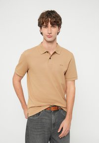 Μπεζ πόλο πουκάμισο με κοντά μανίκια, γιακά και διακριτική λεπτομέρεια λογότυπου. Συνδυασμένο με γκρι τζιν και καφέ ζώνη. Λεία υφή από βαμβάκι.