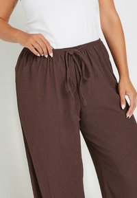 Pantalones anchos de color marrón, texturizados, con una tela ligera y una cinturilla ajustable con cordón, parcialmente mostrados con una mano descansando en la cintura.