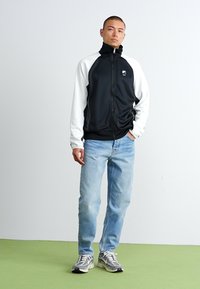 Crna i bijela zip majica s visokim ovratnikom, uparen s svijetloplavim trapericama i sivim tenisicama. Glatka tkanina, sportski kroj, vidljiv detalj s logotipom.