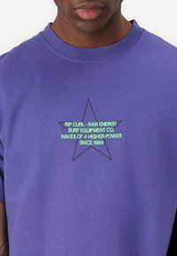 Fialové bavlněné tričko s grafickým motivem zelené a černé hvězdy a textem "Rip Curl - Raw Energy Surf Equipment Co. Od roku 1969."
