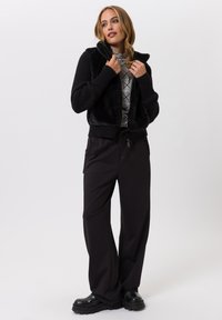 Zwarte faux fur cropped jas met ribbels aan de mouwen, gecombineerd met hoge zwarte broeken. Het model heeft lang haar in losse golven gestyled.