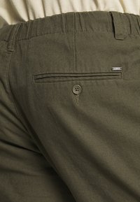 Pantaloni verde oliva realizzati in tessuto testurizzato, con una tasca sul retro con bottone e un'etichetta. La vita è elasticizzata per un maggiore comfort.