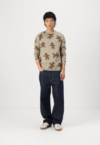 Beige knit sweater featuring brown human figure patterns, paired with loose dark blue denim jeans and white sneakers.