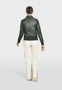 Giacca di pelle verde con un design aderente, polsini e vita a coste. Indossata sopra una camicia chiara e abbinata a pantaloni bianchi.