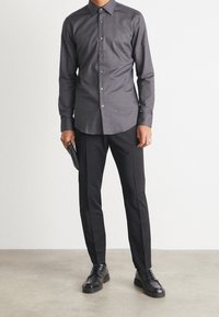 Grijs overhemd met knopen en een standaardkraag, gecombineerd met op maat gemaakte zwarte broek en zwarte leren schoenen, met een zwarte clutch in de hand.