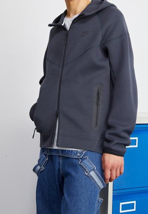 Person trägt einen dunkelgrauen Nike-Zip-Hoodie und blaue Jeans mit Hosenträgern, steht neben blauen und weißen Schubladen.