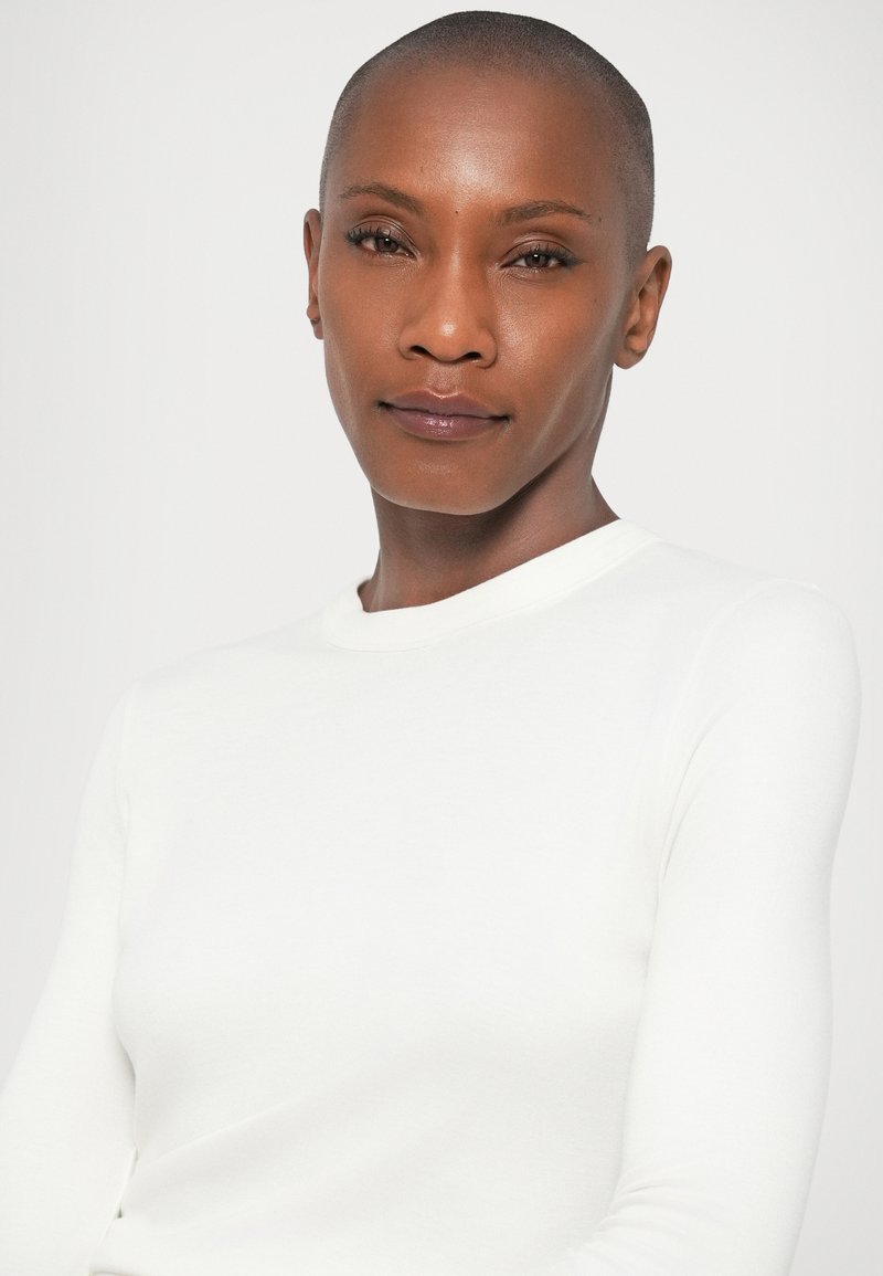 White long-sleeve top with a round neckline, smooth texture, and form-fitting design. Minimalistic aesthetic with no visible patterns or accents.