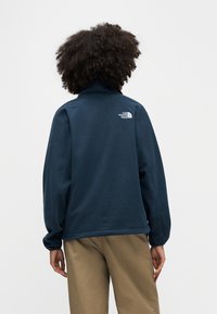 Πουλόβερ από φλις σε ναυτικό μπλε με ψηλό γιακά, με μικρό λευκό λογότυπο στην πλάτη και ελαστικά μανίκια. Λεπτή υφή, άνετη εφαρμογή.