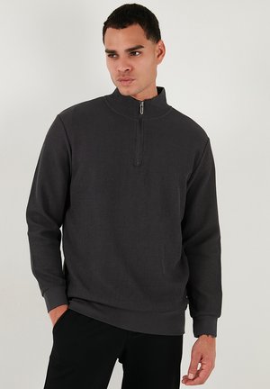 Sudadera de cremallera gris oscuro con textura acanalada, cuello alto y mangas largas. Presenta un diseño sólido y puños y dobladillo elásticos.