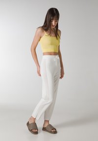 Gelbes, gekürztes Tanktop, weiße Hose mit seitlichem Mesh-Einsatz und braunen Rutsch-Sandalen. Das Material scheint weich und leicht zu sein. Lässig geschnitten.