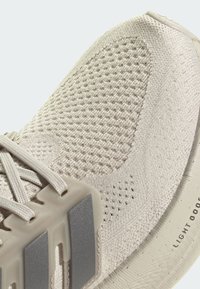 Zapatilla deportiva de punto beige con parte superior texturizada, cordones beige, tres franjas reflectantes y el texto "LIGHT BOOST" en la suela acolchada.