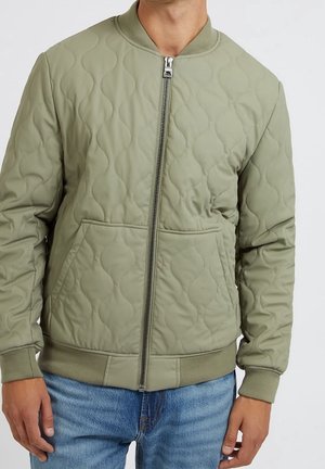 Homme portant une veste bomber matelassée vert clair avec fermeture éclair et poches, associée à un jean bleu, visible du cou aux hanches.