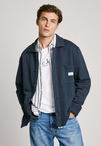 Marinblå jacka med knappar, spetsig krage och framficka, lager över en vit skjorta med blå ränder, tillsammans med ljusblå jeans.