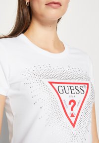 Hvid T-shirt med korte ærmer, der har et rødt triangel-logo omgivet af sølvfarvede rhinsten, med en glat bomuldsstruktur.