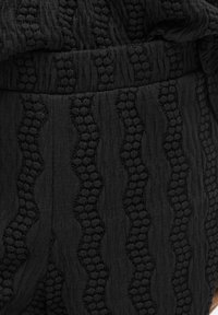 Un tissu noir texturé présente des motifs ondulés, avec une broderie en relief qui crée de la profondeur. La surface lisse complète les détails délicats.