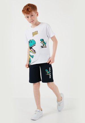 Punapäine poiss kannab valgeid tossusid, musti lühikesi pükse dinosaurusetrükiga ja valget T-särki, millel on rula sõitev dinosaurus ja tekst.