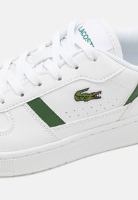 Hvite lær sneakers med grønne detaljer, med perforeringer, flate hvite lisser, og en liten brodert krokodillelogo. Flat gummisåle.