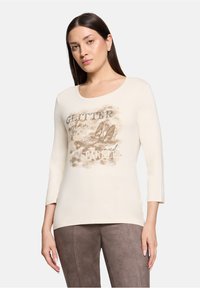 Cremefarbenes Langarmshirt mit einem grafischen Druck, der glitzernde Akzente und stilisierte Schuhillustrationen zeigt. Glatte Stofftextur.