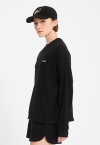 Jeune personne vêtue d'un t-shirt à manches longues noir et d'un short, portant une casquette noire avec un motif de scorpion, debout de profil sur un fond blanc.