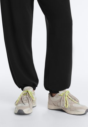 Picioare purtând pantaloni negri de tip jogger și adidași bej cu șireturi galben neon, stând pe o suprafață albă.