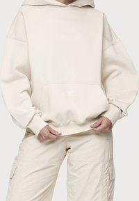 Krämfärgad oversized hoodie i mjukt material med en känguruficka och ribbade ärmslut. Texten på fickan lyder "Skapa det med mig."