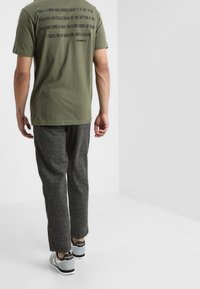 T-shirt de algodón verde oliva con texto negro en la parte posterior, combinada con pantalones texturizados grises y zapatillas grises con acentos negros.