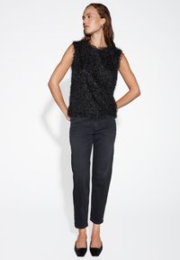 Zwarte mouwloze top van pluizig textuurmateriaal, gecombineerd met hoge taille donkerblauwe jeans en zwartepuntige flats. Eenvoudige achtergrond.